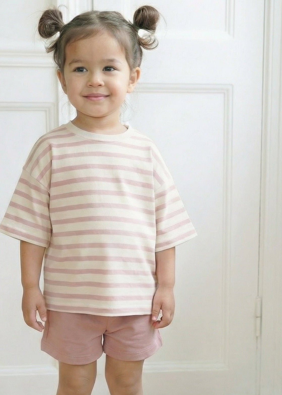 Young girl wearing a striped shirt and shorts standing in a room with white walls.