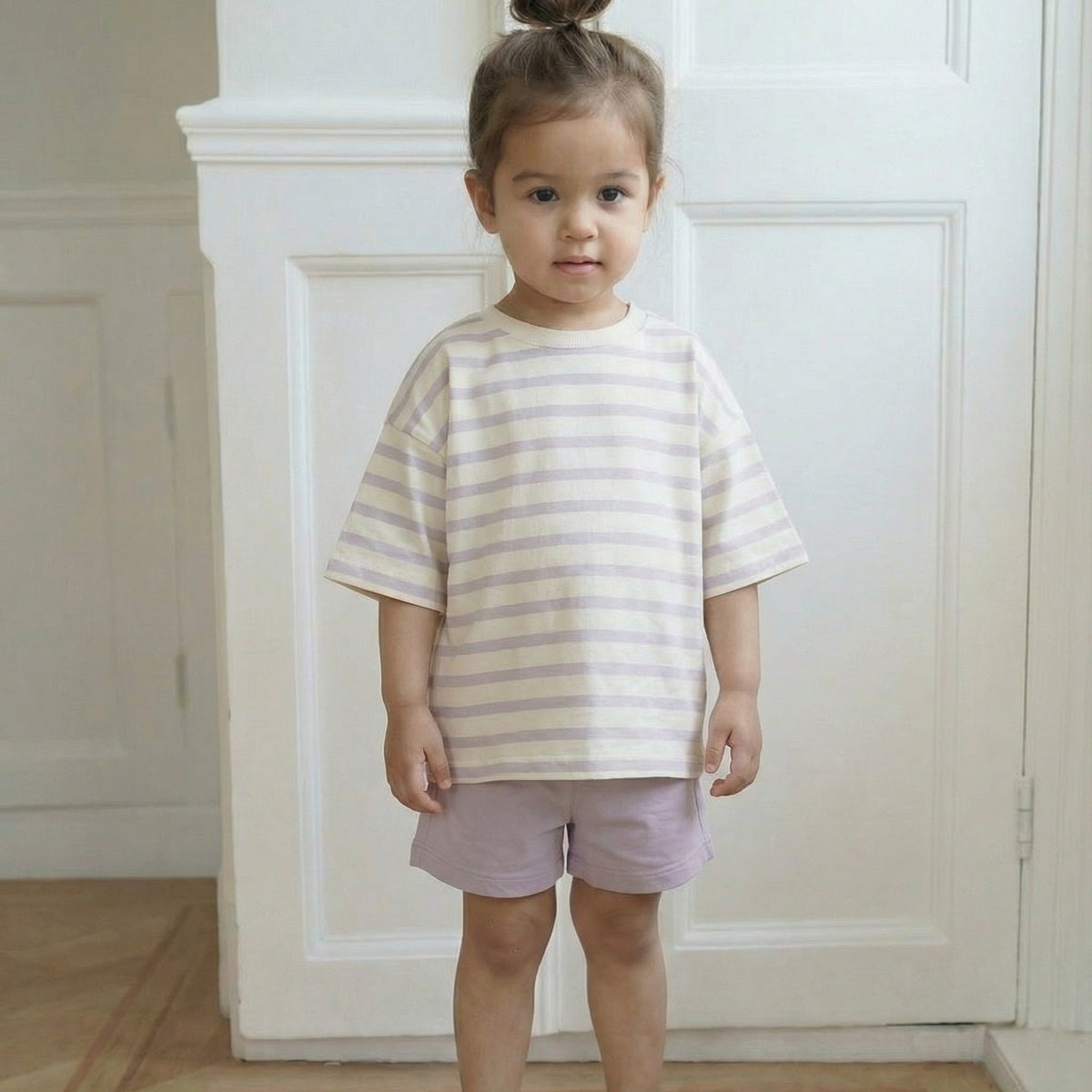 Child wearing a striped shirt and shorts standing in a room with white walls and wooden floor.
