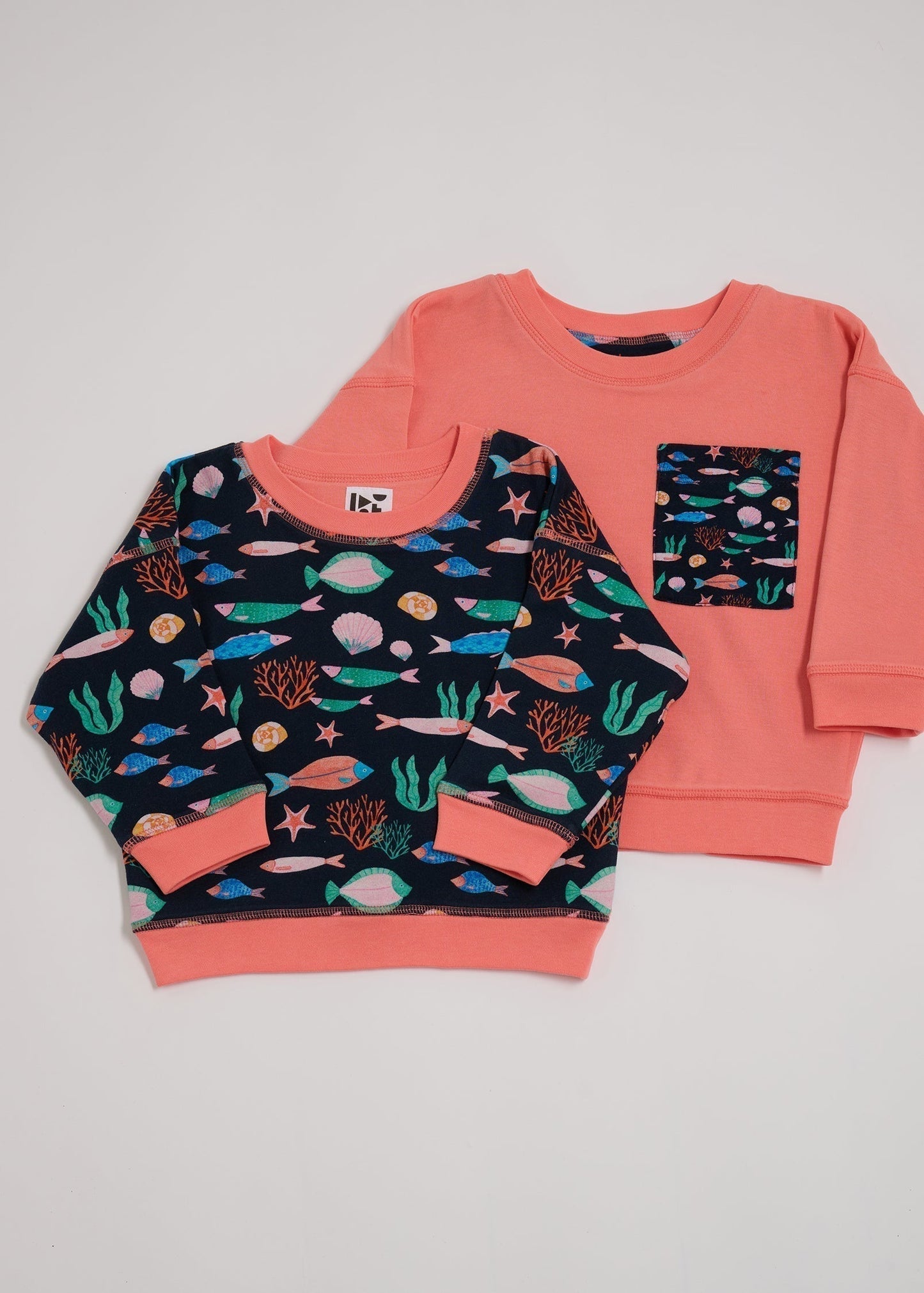 Shoulder to shoulder comparison of both sides of a reversible sweatshirt. One featuring an all over vibrant navy fish print with contrast coral neck and sleeve trim. The other coral with an oversized vibrant navy fish print chest pocket.