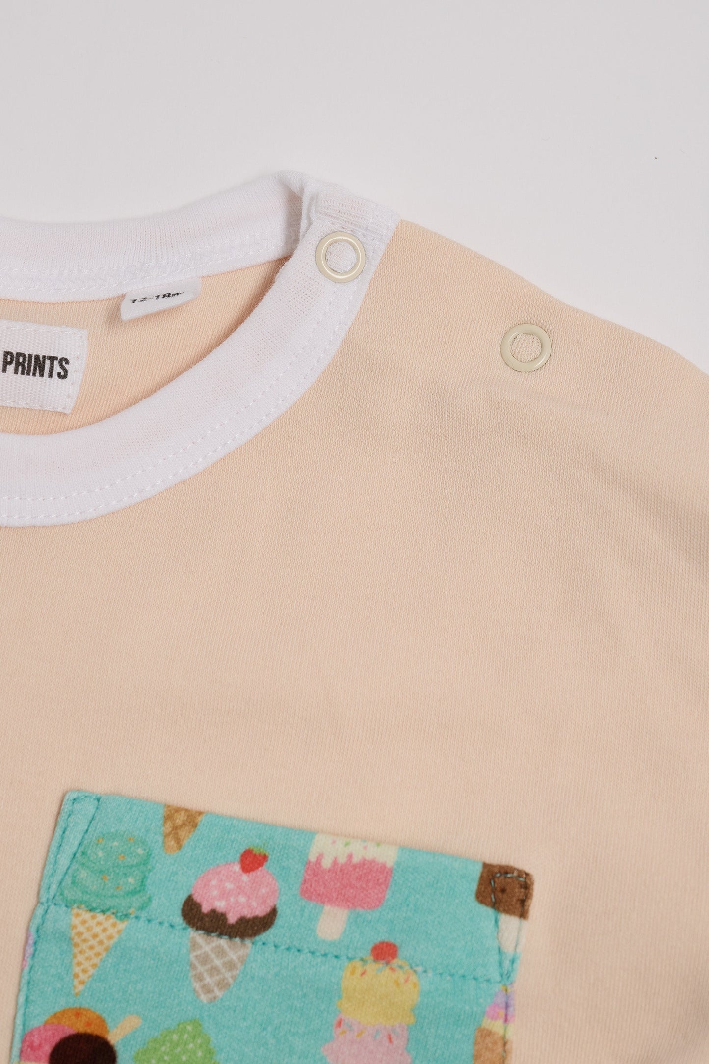 Close-up of neck poppers on vanilla coloured t-shirt with bright ice cream print square chest pocket and white neck trim.