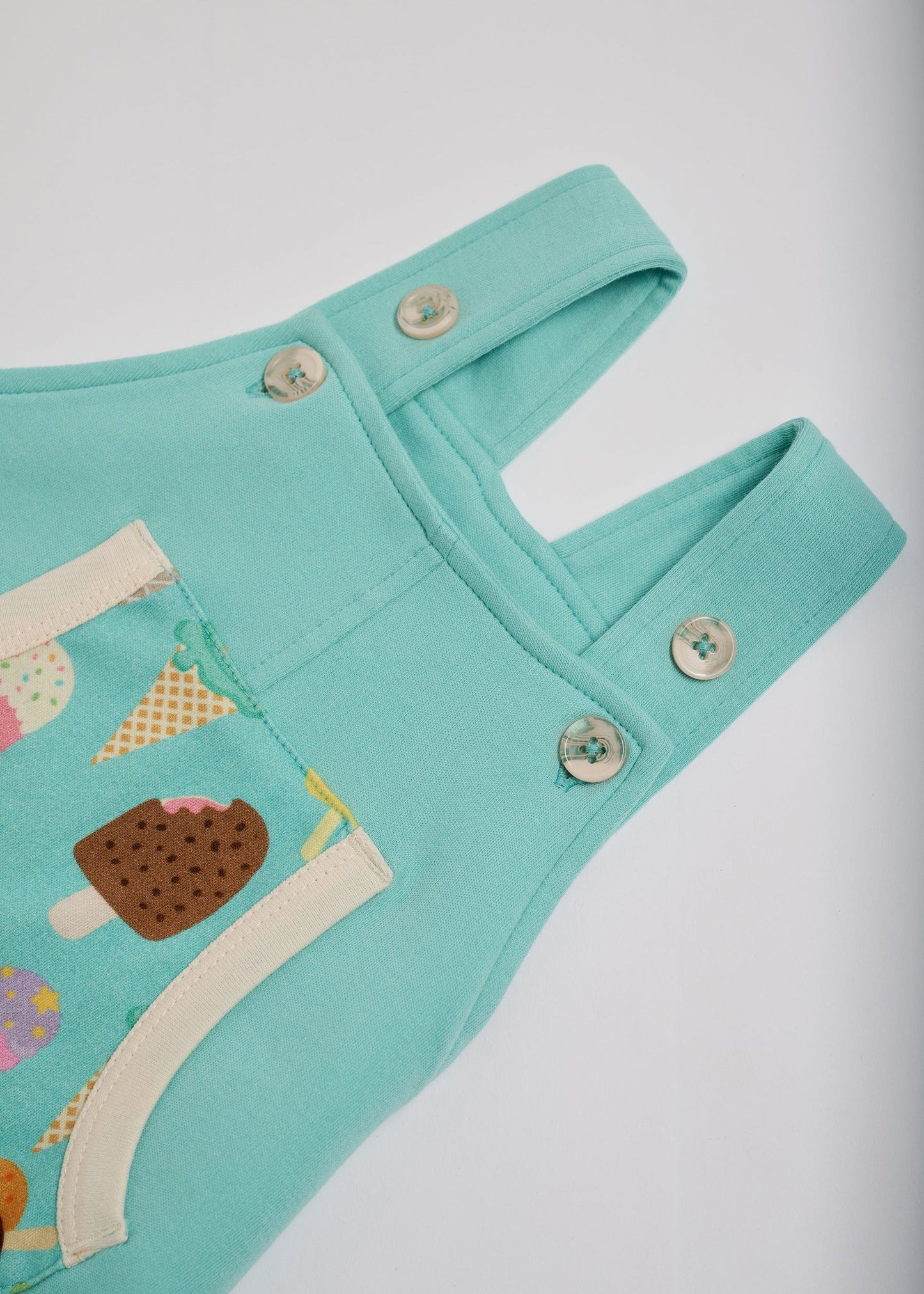 High setting on adjustable straps on turquoise short dungarees with bright ice cream print pocket, vanilla buttons and vanilla trim.