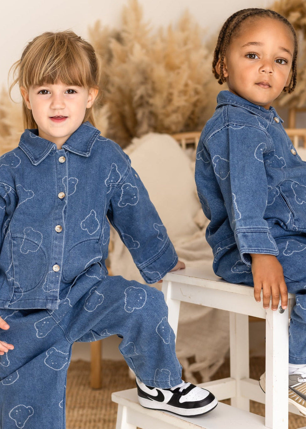 Two children wearing matching denim outfits sitting on a small white stool.