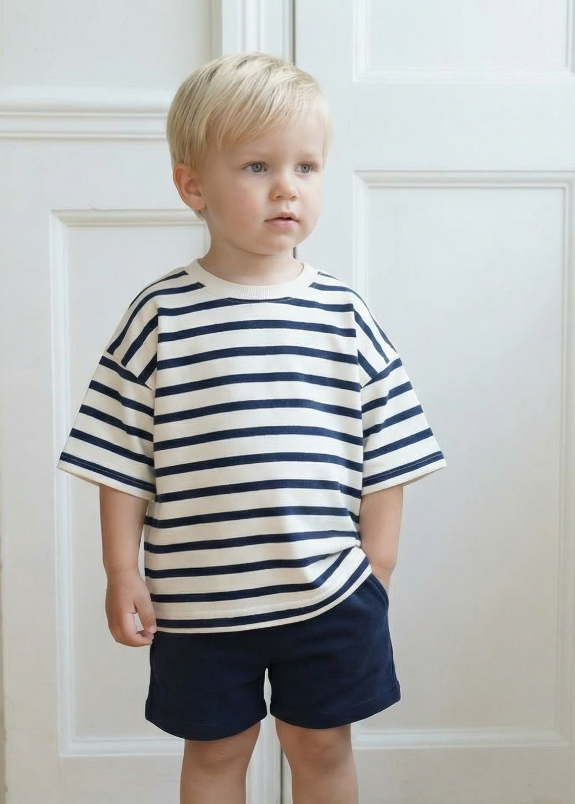 Child wearing a striped shirt and shorts standing in a room with white walls and wooden floor.
