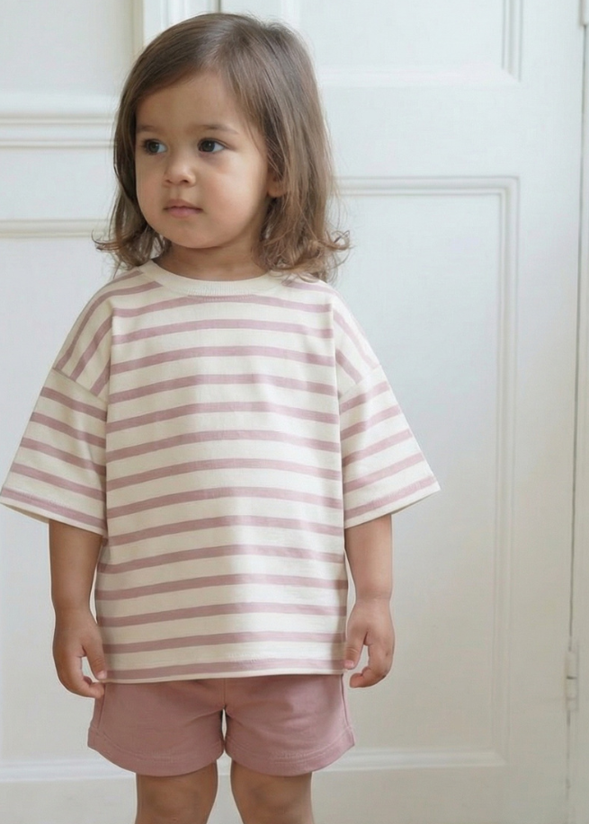 Child wearing a striped shirt and shorts standing in a room with white walls and wooden floor.