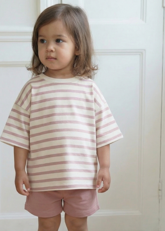 Child wearing a striped shirt and shorts standing in a room with white walls and wooden floor.