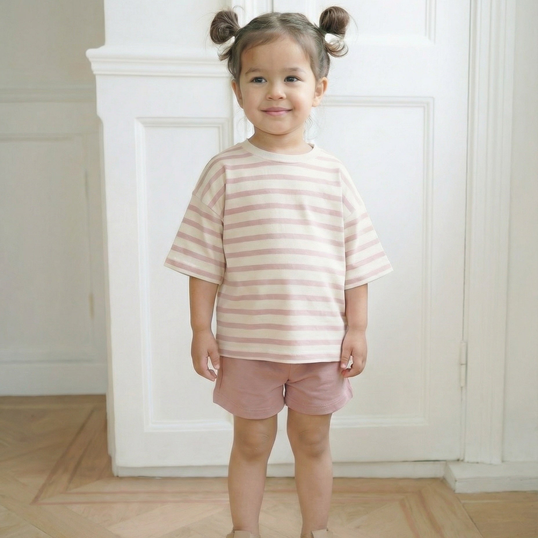 Young girl wearing a striped shirt and shorts standing in a room with white walls.