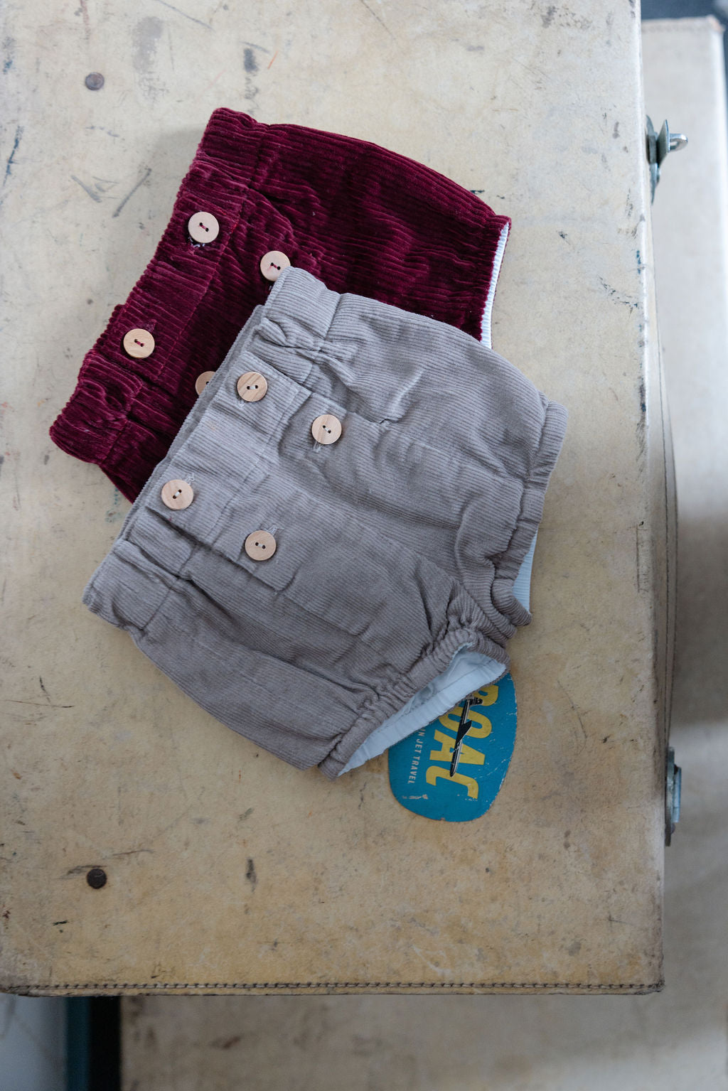 Two pairs of children's shorts, one maroon and one gray, on a metal surface.