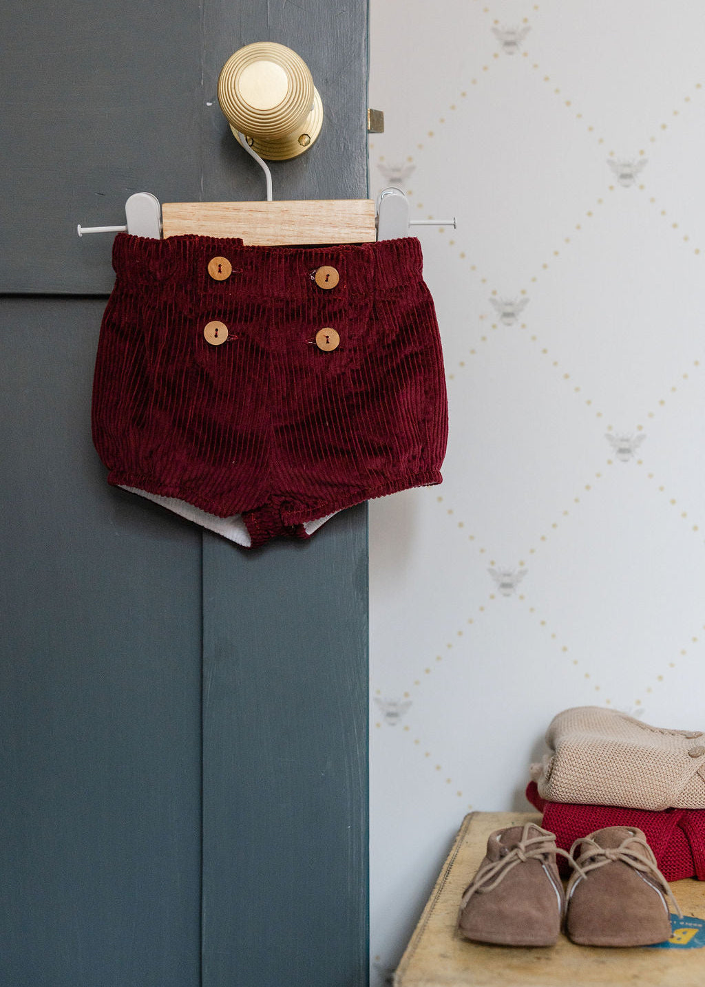 Red corduroy shorts with gold buttons hanging on a door next to a wall with a light fixture.