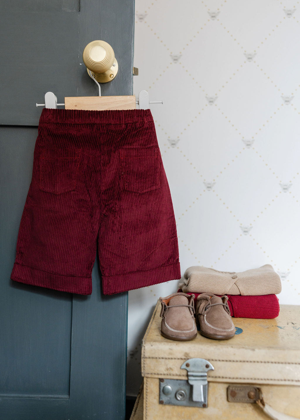 Red corduroy pants hanging on a hook with a suitcase and shoes below.