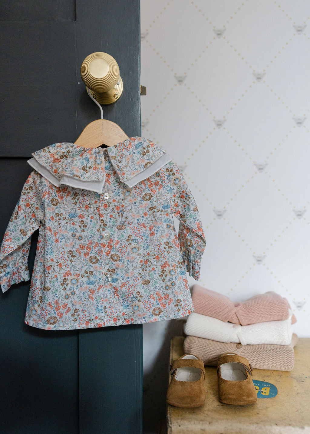 Floral-patterned children's coat on a hanger against a wall with shoes and folded clothes below.