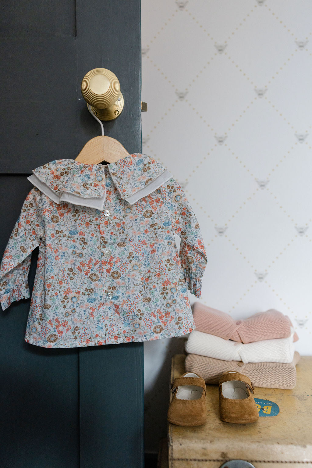 Floral-patterned children's coat on a hanger against a wall with shoes and folded clothes below.