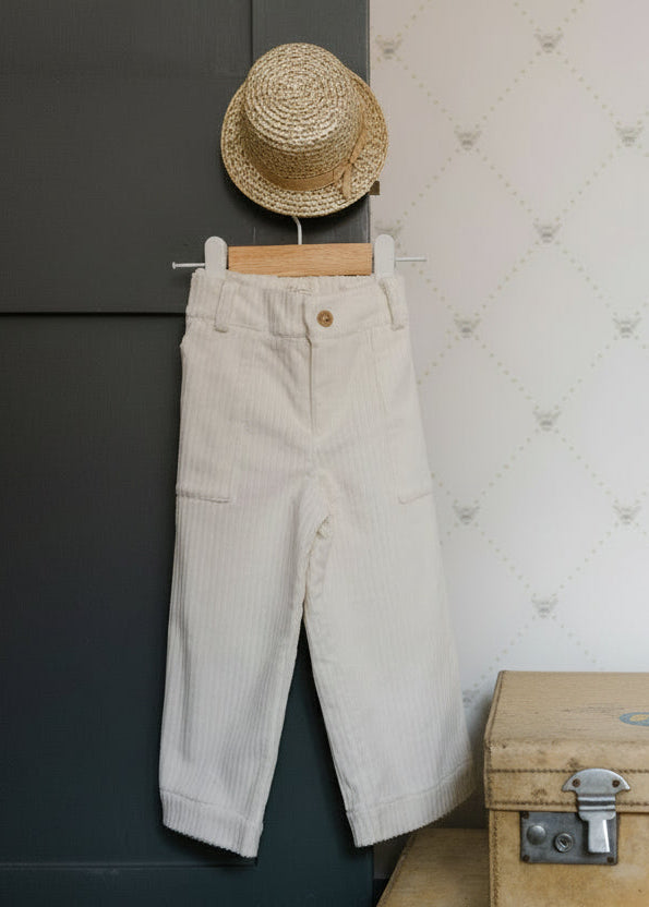 White pants hanging on a hook next to a stack of folded clothes and a vintage suitcase against a white wall.