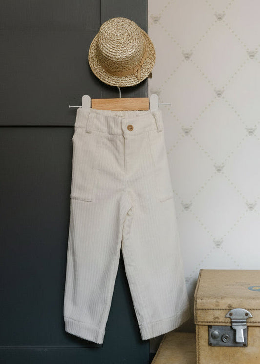 White pants hanging on a hook next to a stack of folded clothes and a vintage suitcase against a white wall.