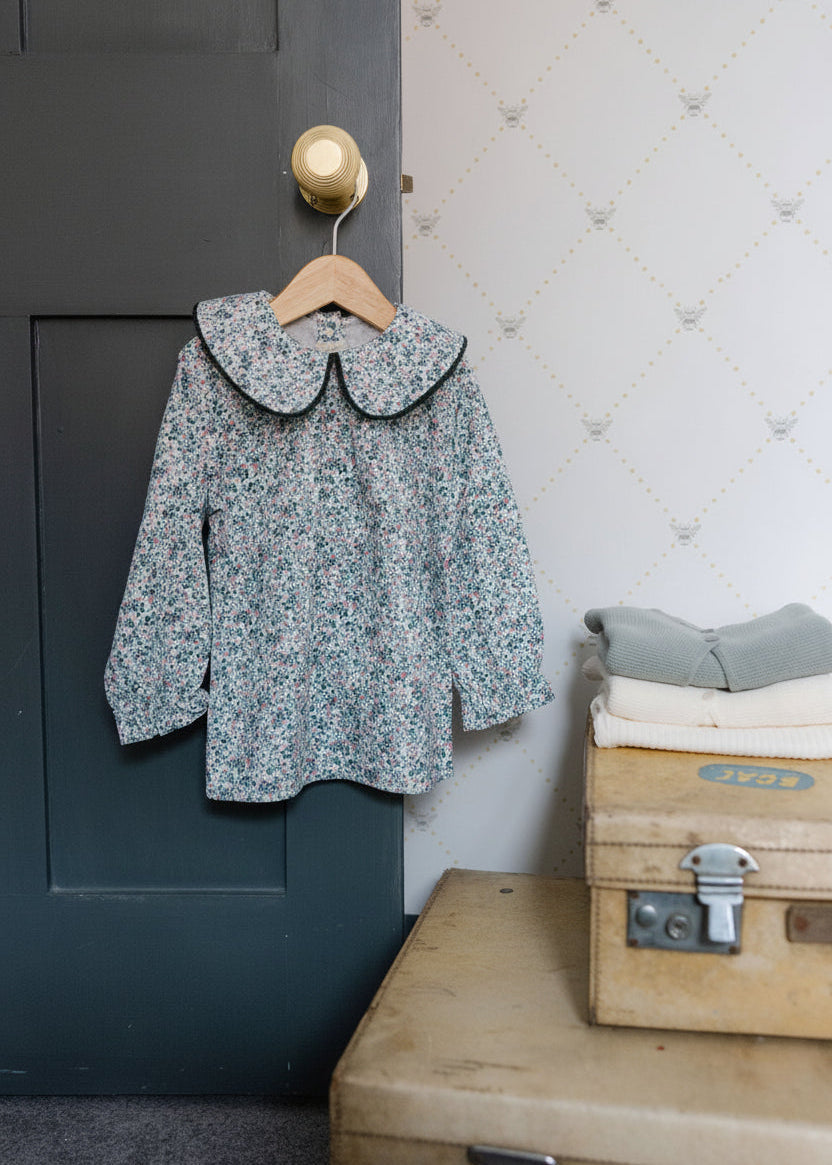 Floral blouse with black collar on a hanger against a textured wall.