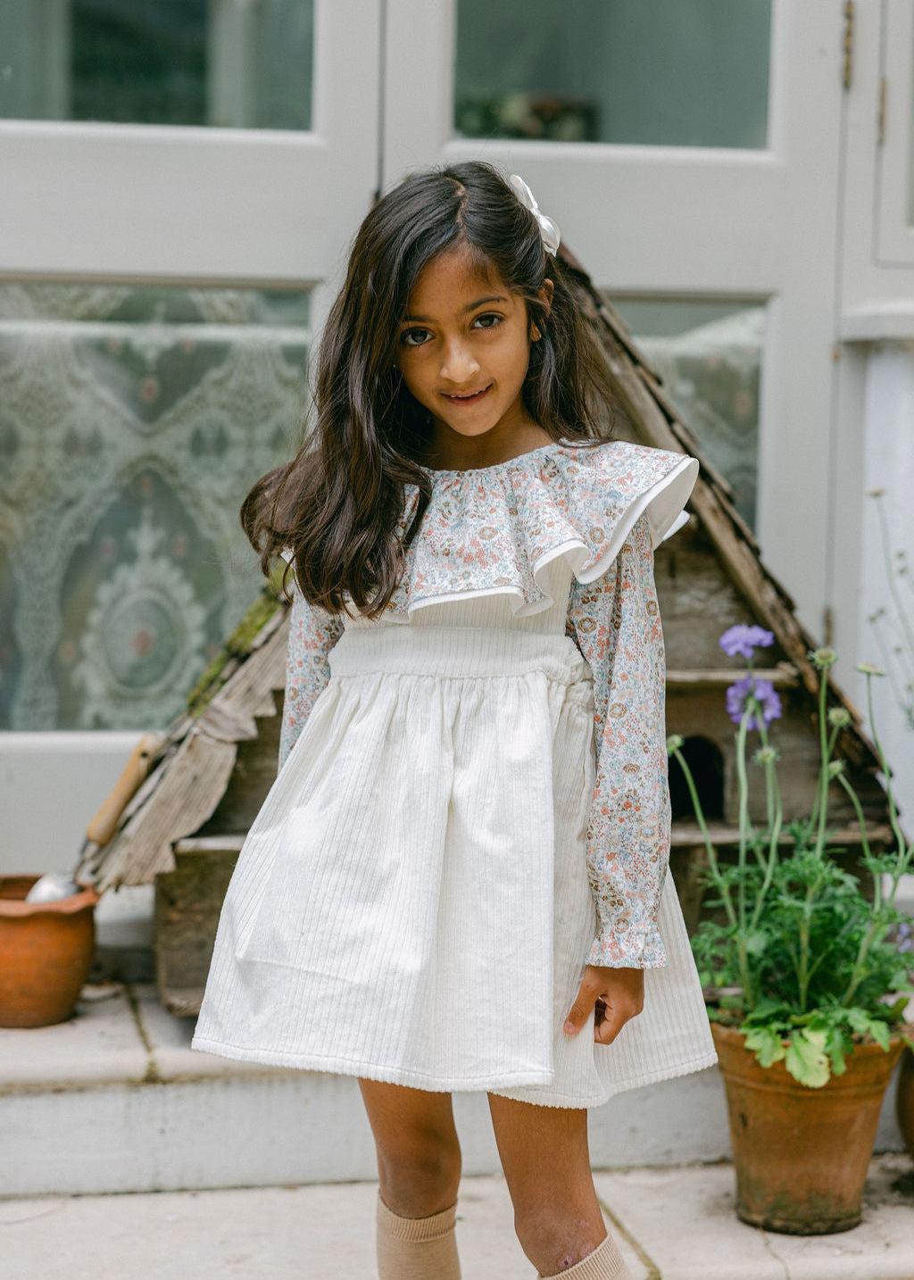 Young girl in a white dress standing outdoors with plants and a wooden structure in the background