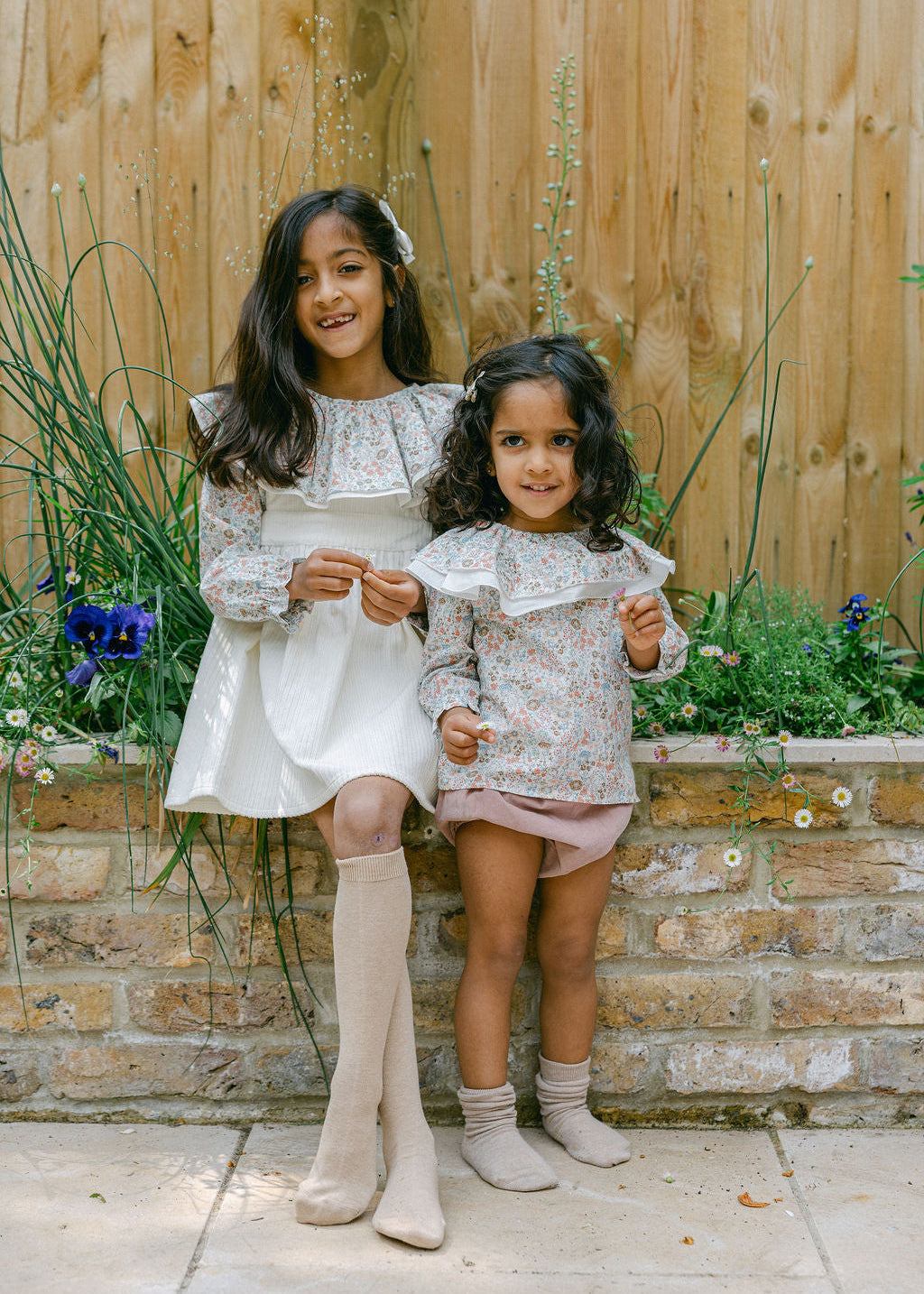 Two young girls in matching outfits standing outdoors against a wooden fence and stone wall.