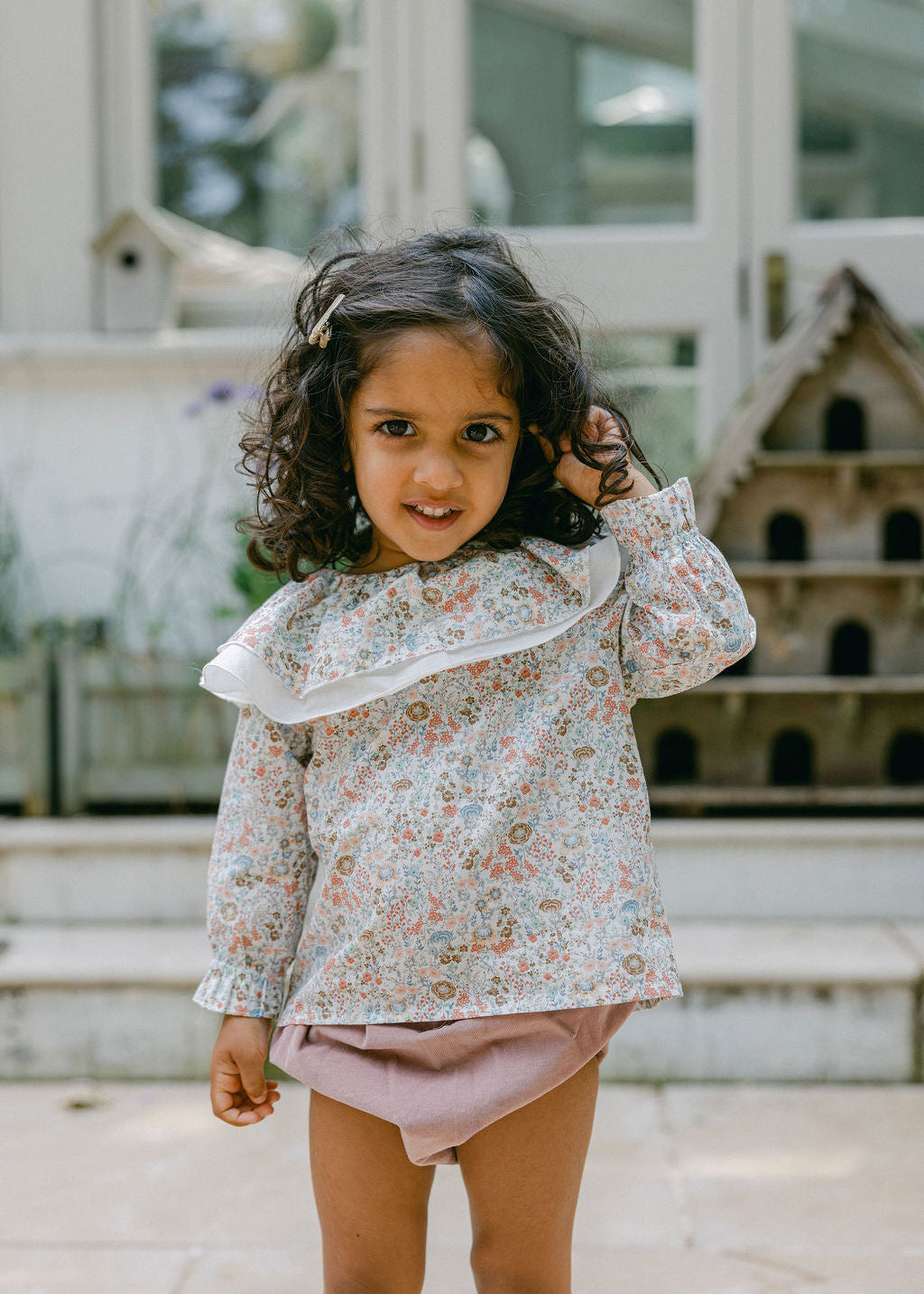 Young girl wearing a floral blouse and pink shorts standing outdoors.