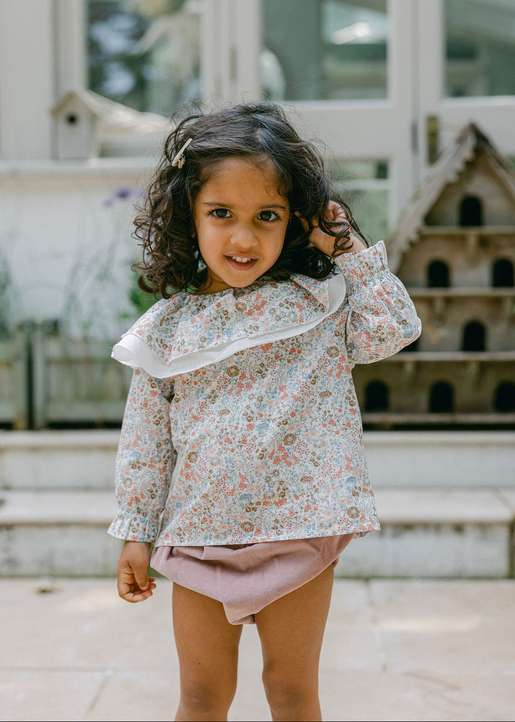 Young girl wearing a floral blouse and pink shorts standing outdoors.