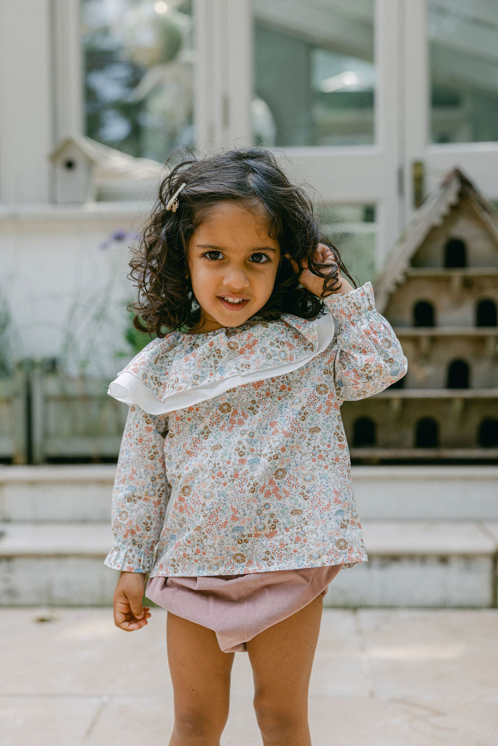Young girl wearing a floral blouse and pink shorts standing outdoors.