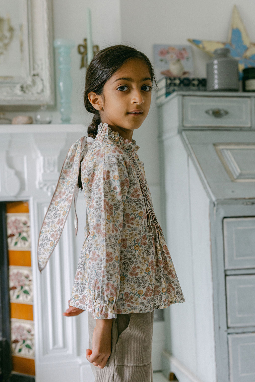 Young girl wearing a floral blouse standing in a room with white cabinets and decorative items.