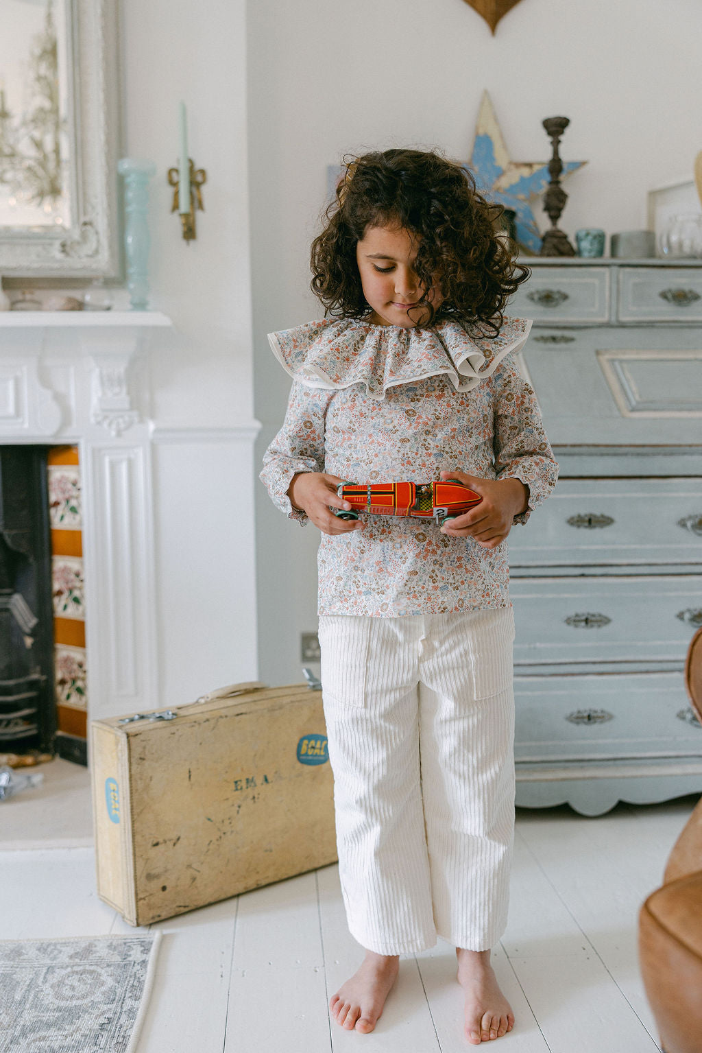Child holding a toy in a room with a fireplace and dresser.