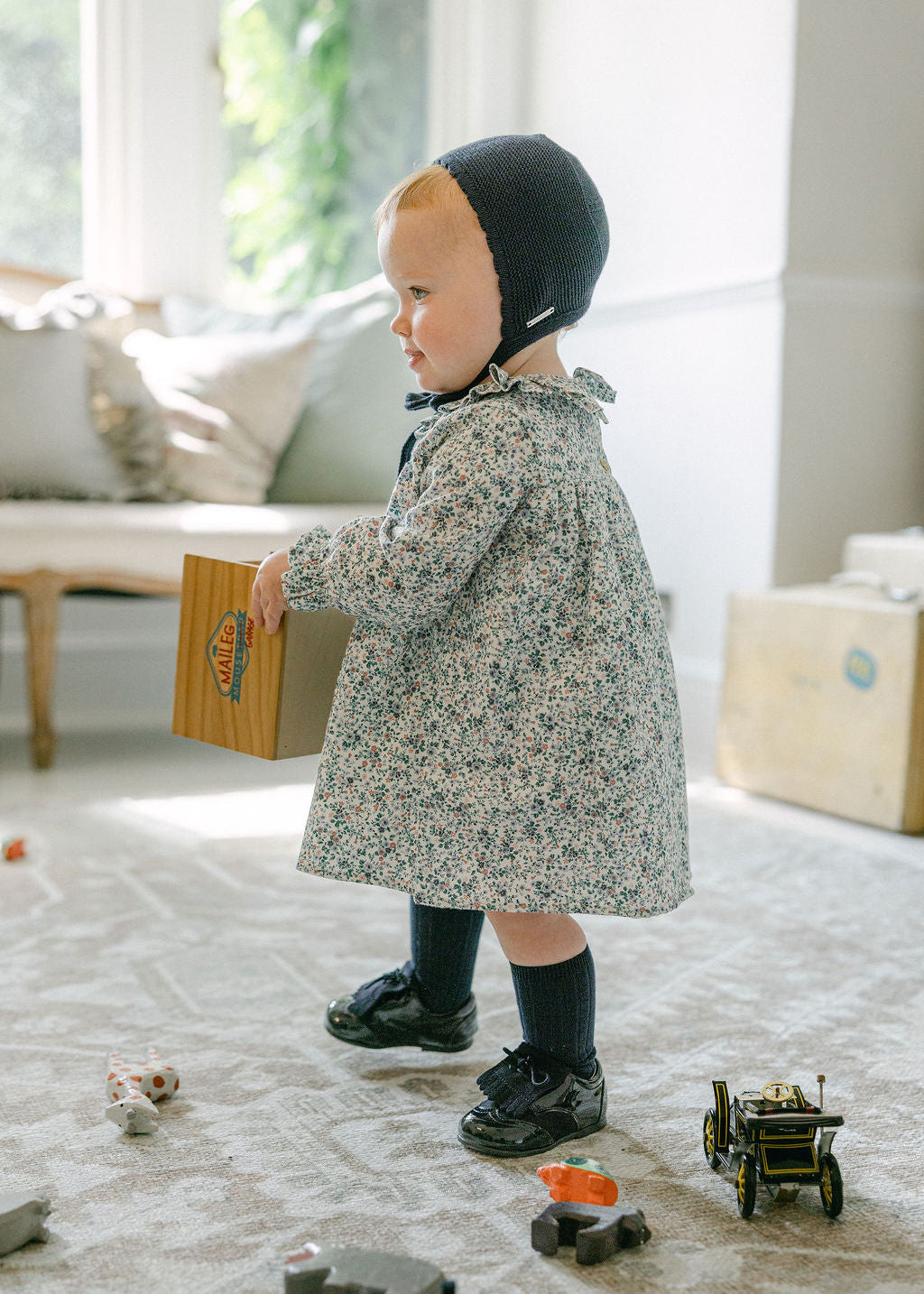 Child in a floral coat and black bonnet playing with toys on a carpeted floor.