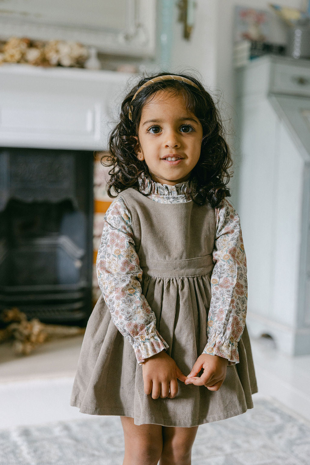 Young girl in a beige dress with floral sleeves standing in a home setting.