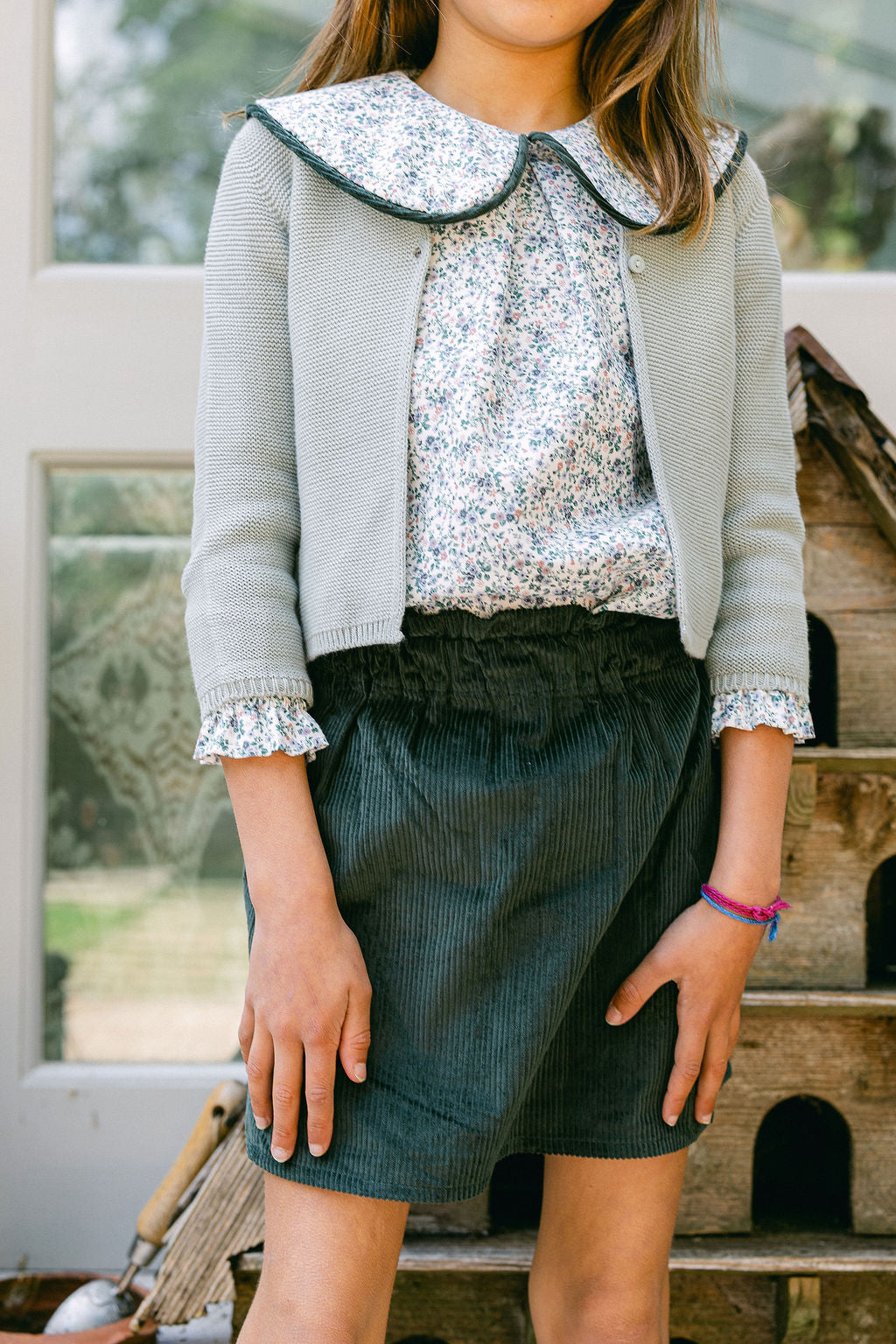 Person wearing a sequin top, gray cardigan, and green skirt sitting on a wooden bench.