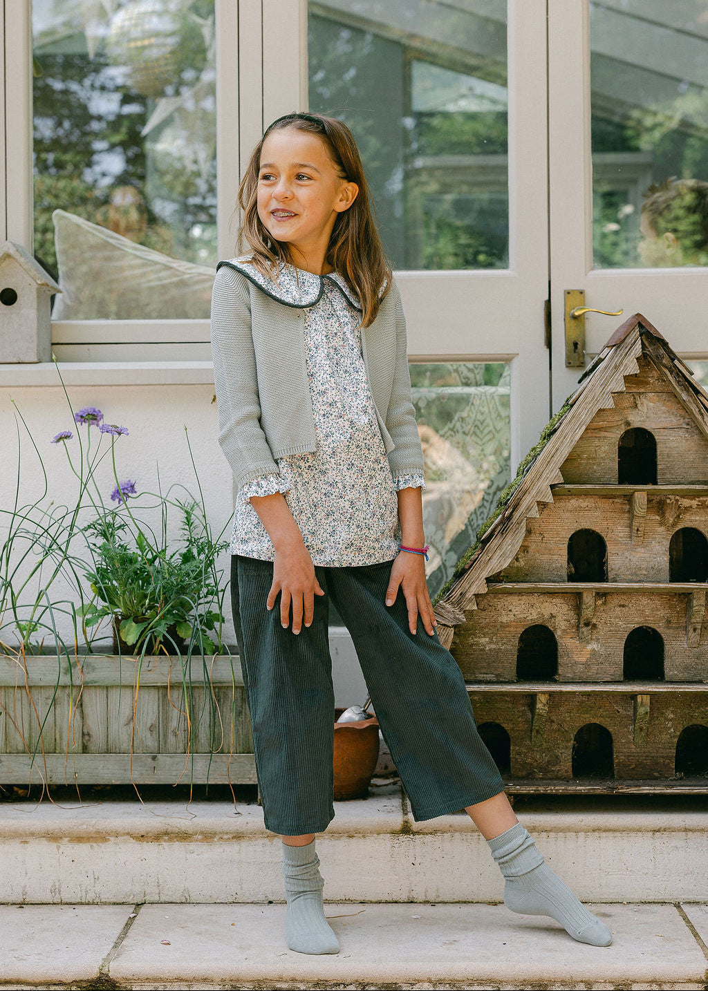 Young girl standing outdoors in front of a greenhouse, wearing a light gray jacket and dark green pants.