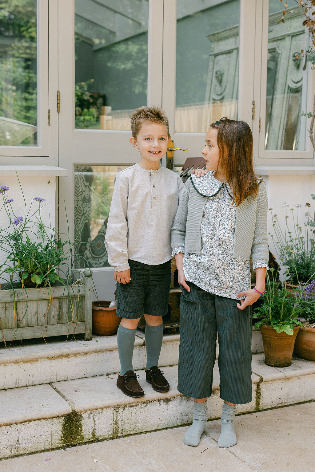 Two children standing on a porch with plants and a house in the background