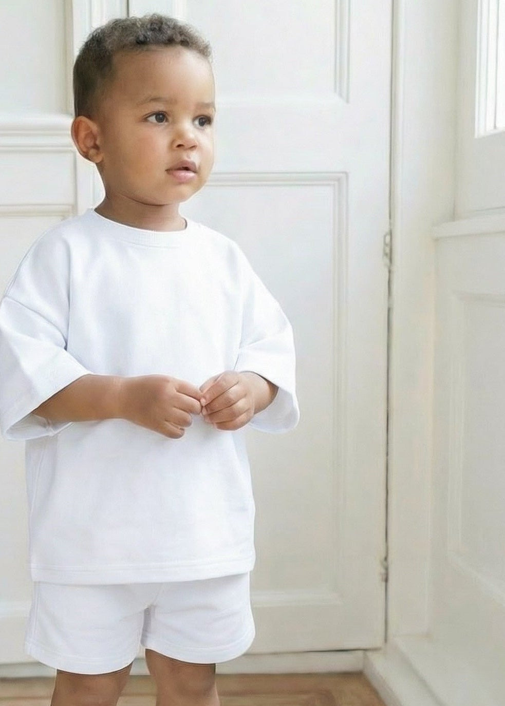 Child wearing a white outfit standing in a bright room with wooden flooring.