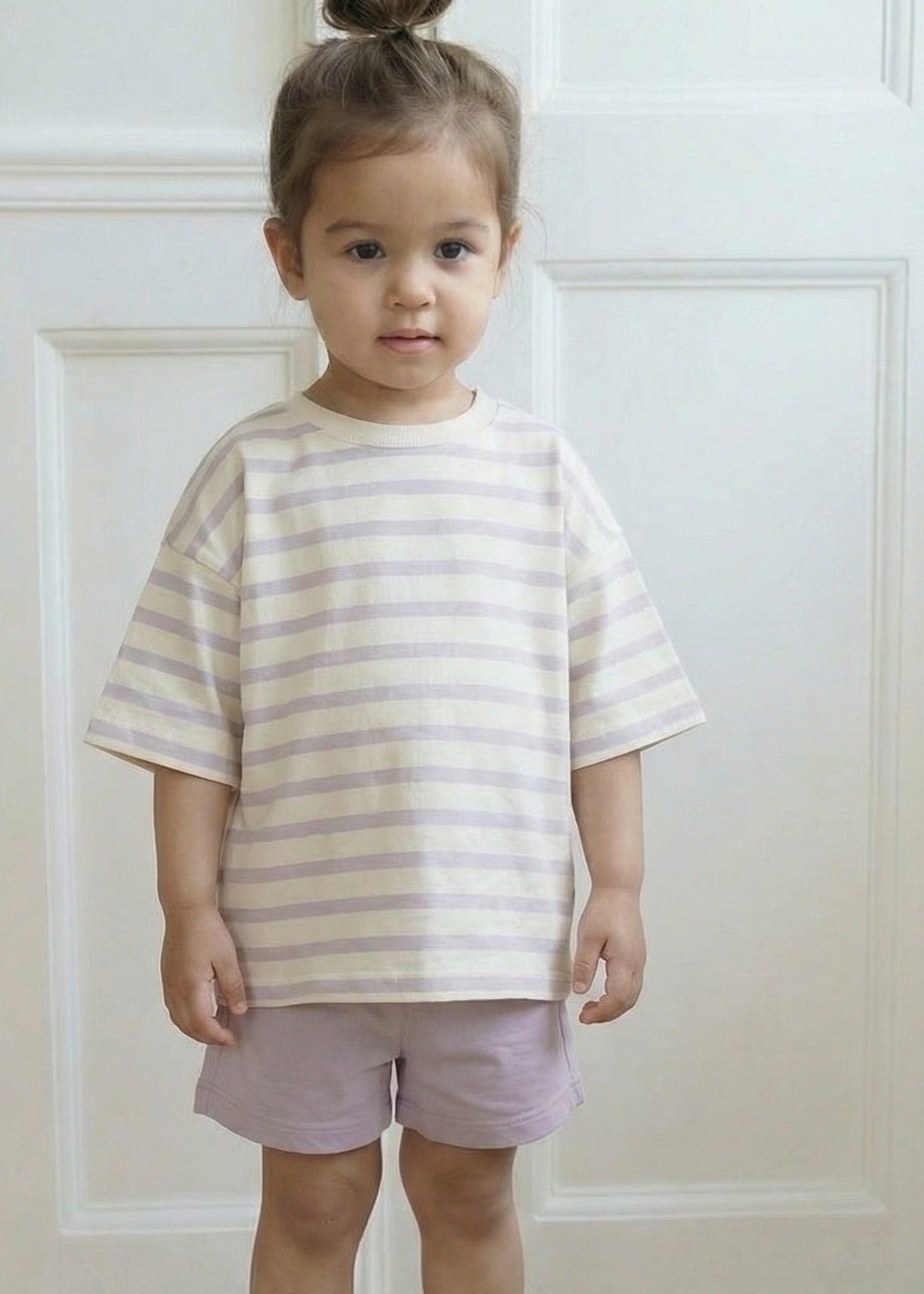 Child wearing a striped shirt and shorts standing in a room with white walls and wooden floor.