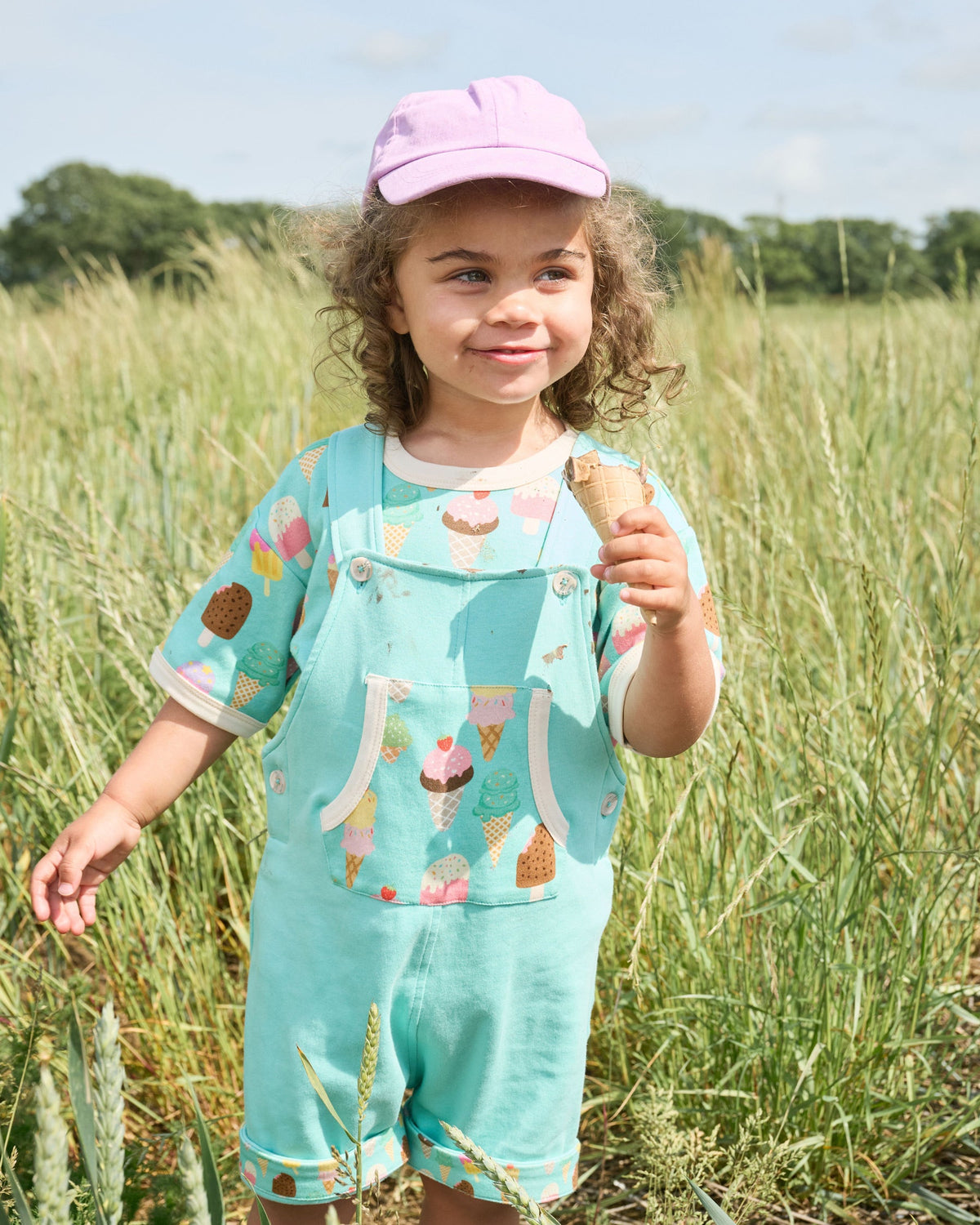 Young girl enjoying an ice-cream in a field wearing a bright ice cream print t-shirt and short turquoise dungarees with bright ice cream print pocket and turn-ups, vanilla buttons and vanilla trim.