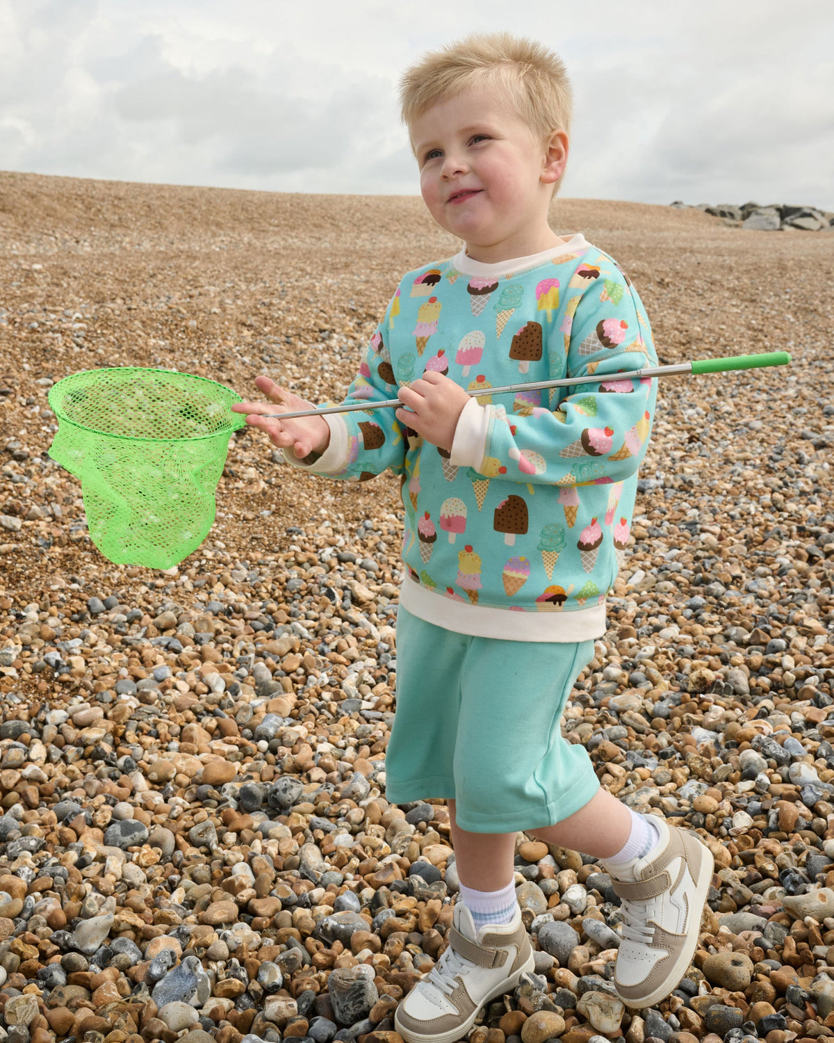 Young smiling boy with a fishing net at the beach wearing a Front of sweatshirt featuring an all over turquoise ice cream print with contrast vanilla neck and sleeve trim over matching turquoise short dungarees.