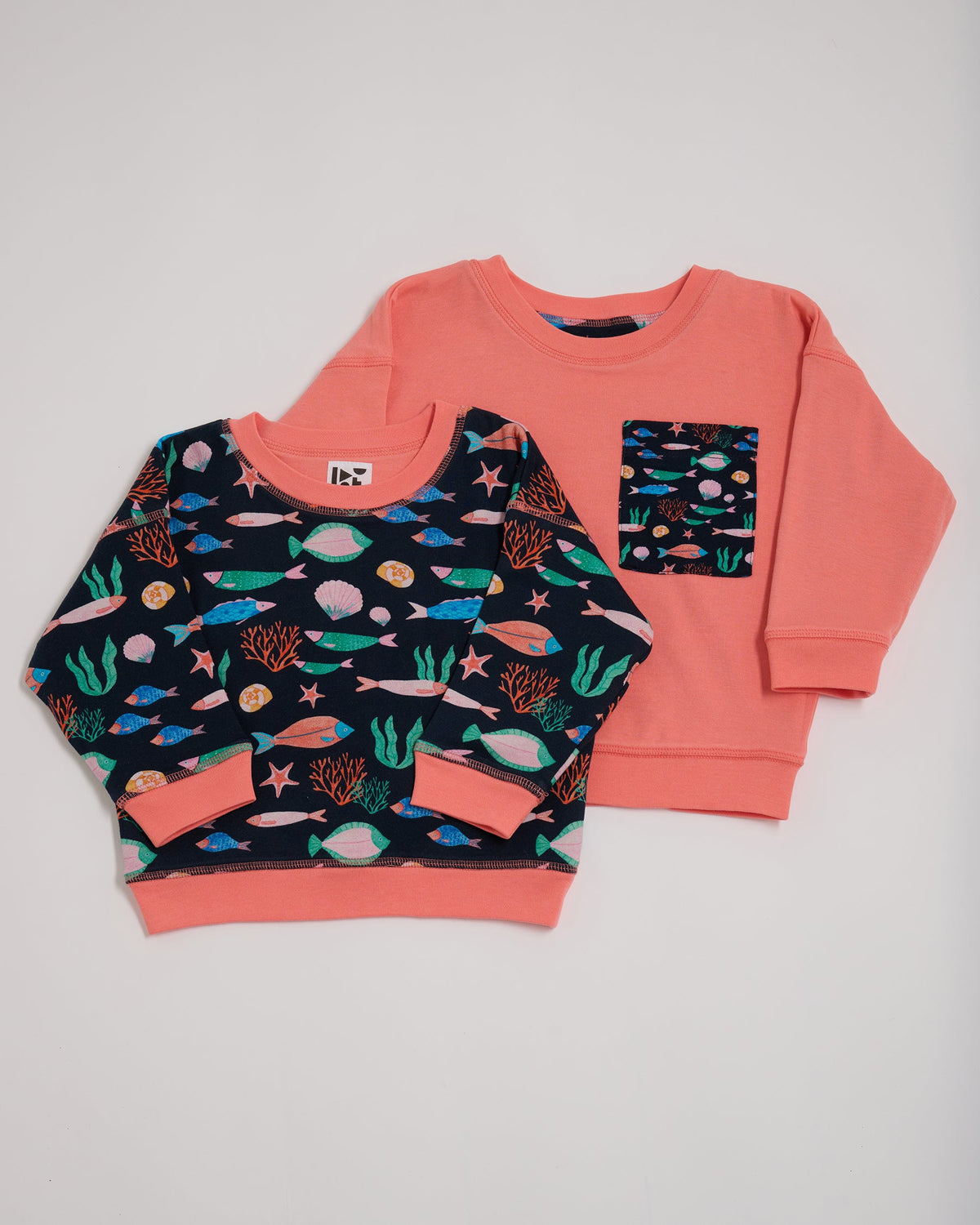 Shoulder to shoulder comparison of both sides of a reversible sweatshirt. One featuring an all over vibrant navy fish print with contrast coral neck and sleeve trim. The other coral with an oversized vibrant navy fish print chest pocket.