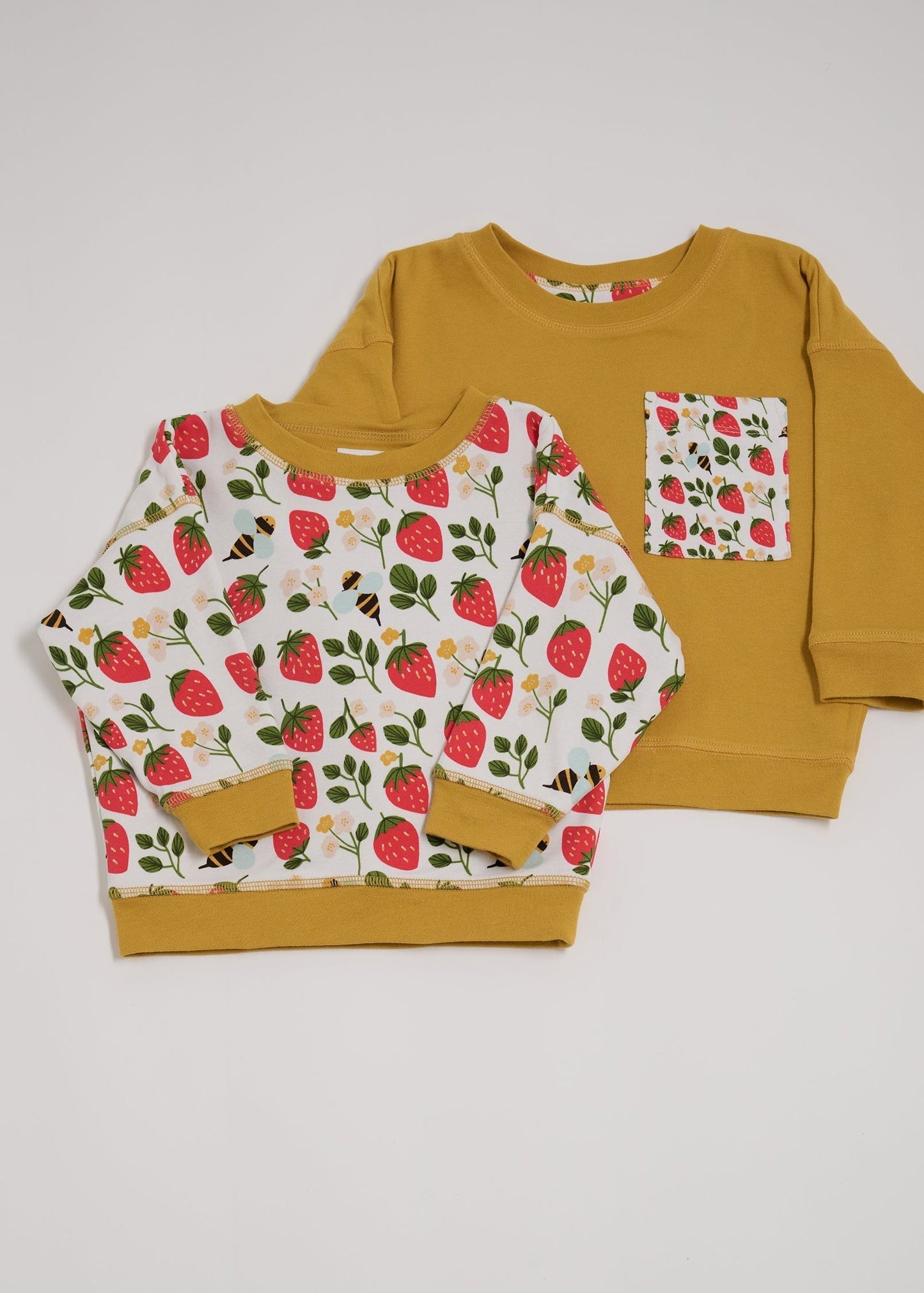 Shoulder to shoulder comparison of both sides of a reversible sweatshirt. One featuring an all over strawberry and bees print with contrast honey neck and sleeve trim. The other honey with an oversized strawberry and bees print chest pocket.
