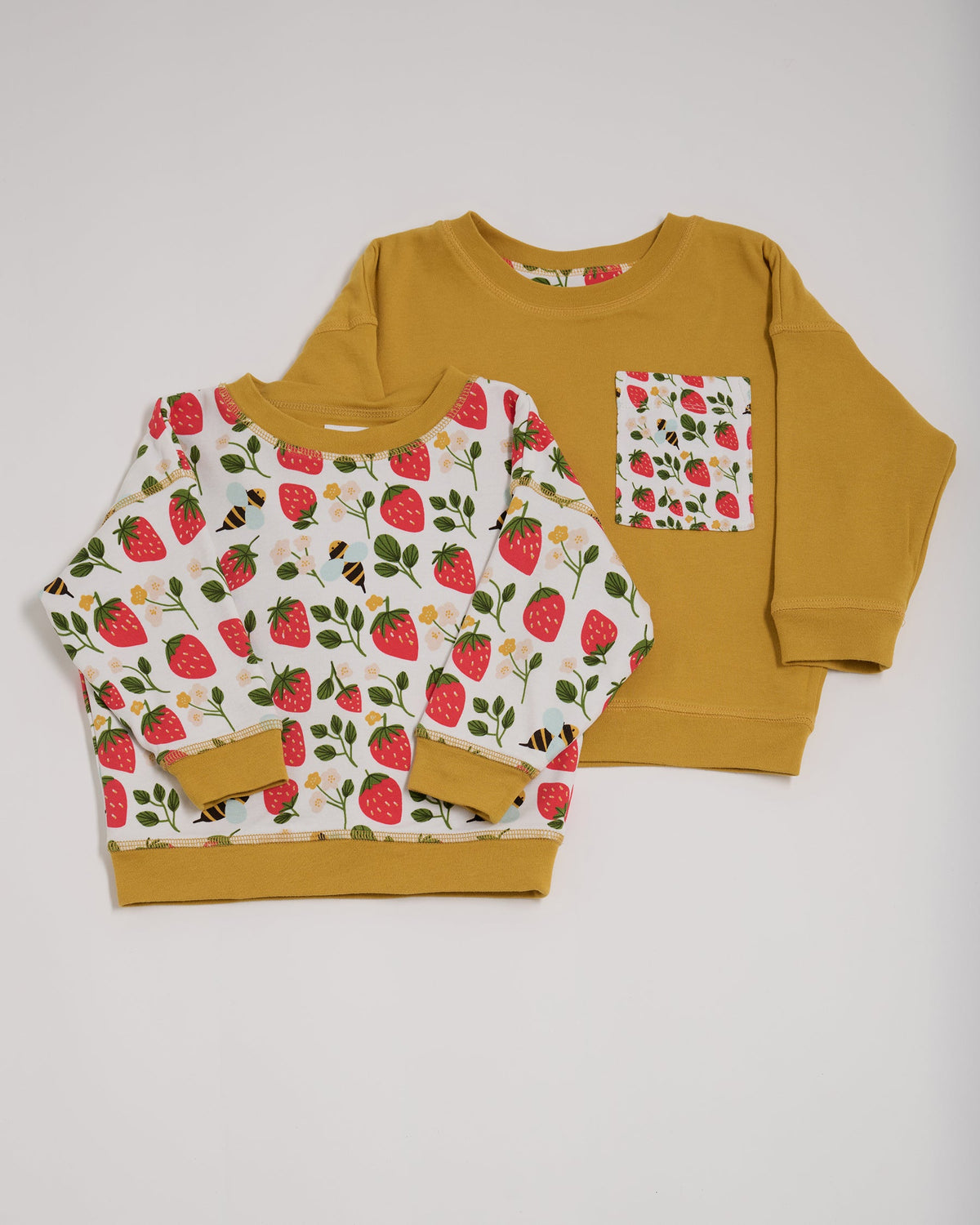 Shoulder to shoulder comparison of both sides of a reversible sweatshirt. One featuring an all over strawberry and bees print with contrast honey neck and sleeve trim. The other honey with an oversized strawberry and bees print chest pocket.