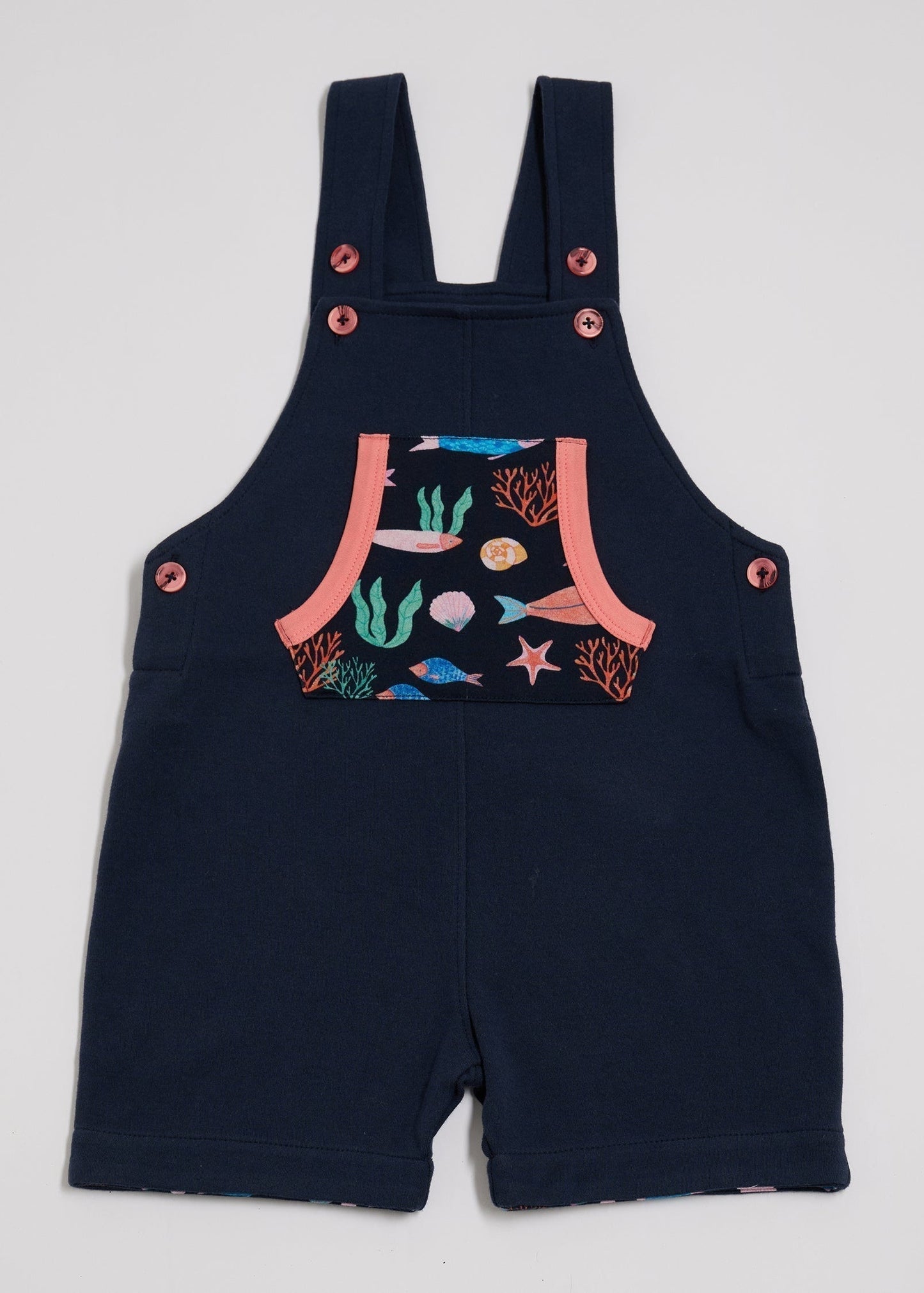 Front of navy short dungarees with vibrant fish print pocket, coral buttons and coral trim.