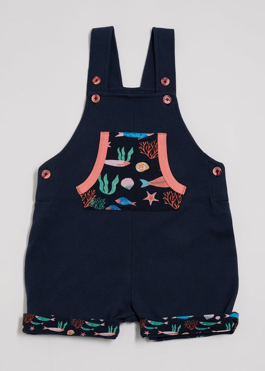 Front view of short dungarees with vibrant fish print pocket and turn-ups, adjustable height straps, coral buttons and coral trim.