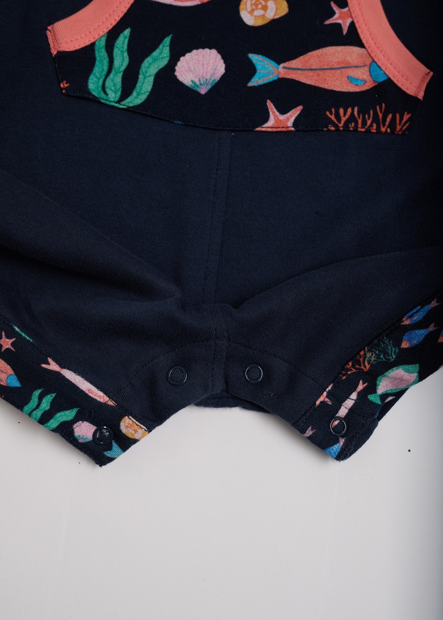Close-up of crotch poppers on navy short dungarees with vibrant fish print pocket and turn-ups and a coral trim.