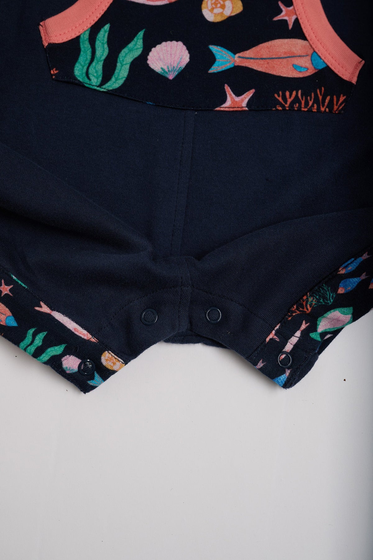 Close-up of crotch poppers on navy short dungarees with vibrant fish print pocket and turn-ups and a coral trim.