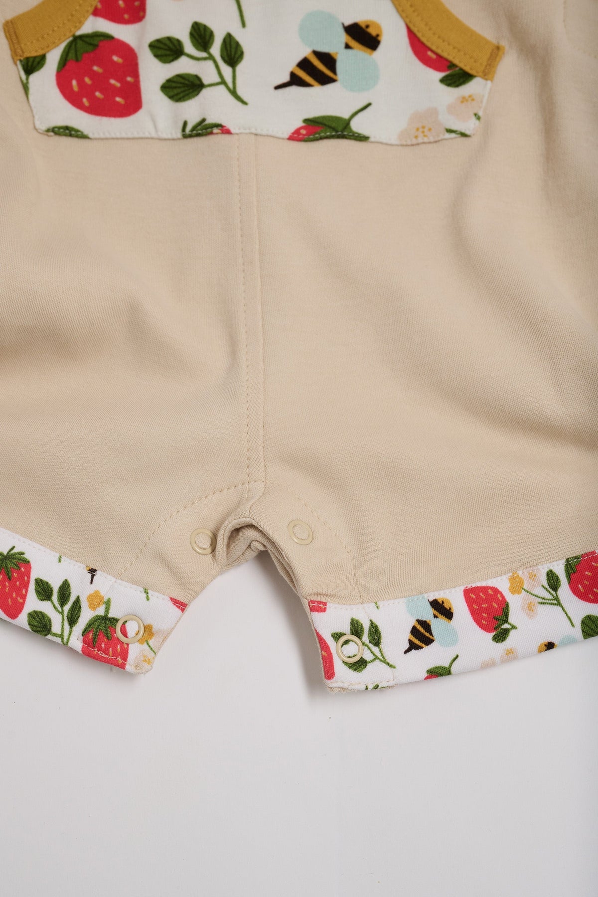 Close-up of crotch poppers on beige short dungarees with fun strawberry and bees print pocket and turn-ups and a honey trim.