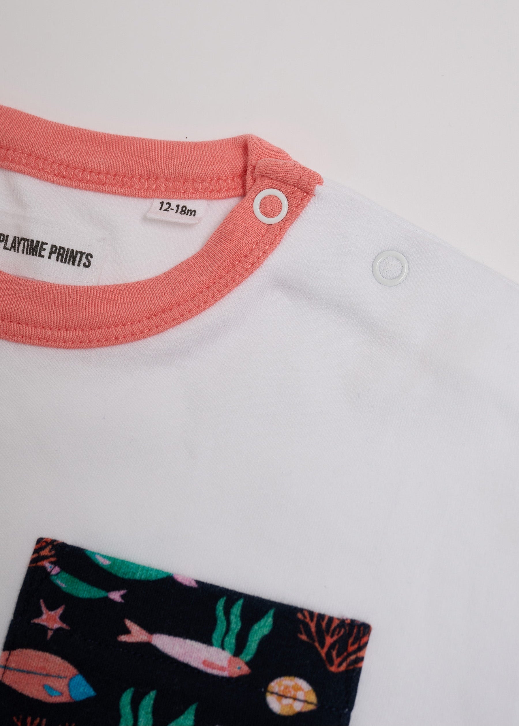 Close-up of neck poppers on white t-shirt with vibrant fish print chest pocket and coral neck trim.