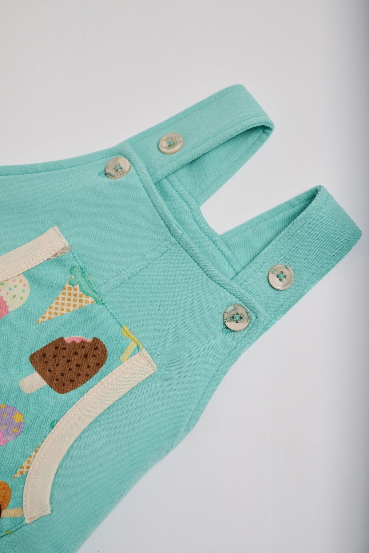 High setting on adjustable straps on turquoise short dungarees with bright ice cream print pocket, vanilla buttons and vanilla trim.