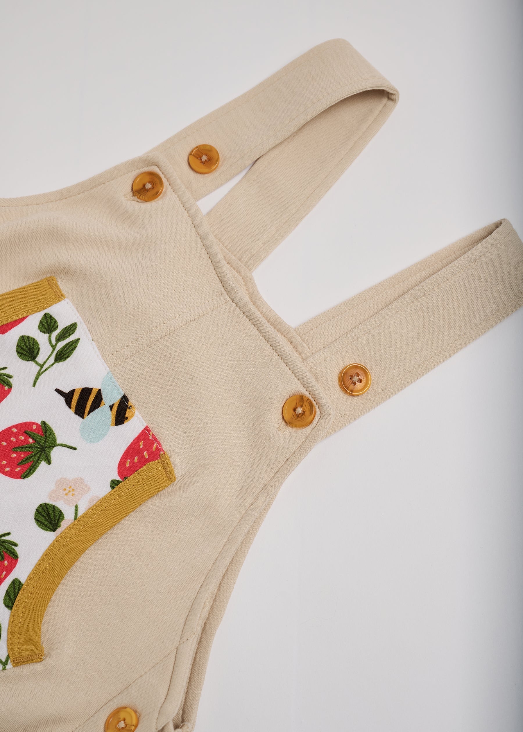 Close-up of adjustable straps set to high setting on beige short dungarees with fun strawberry and bees print and honey buttons and honey trim.