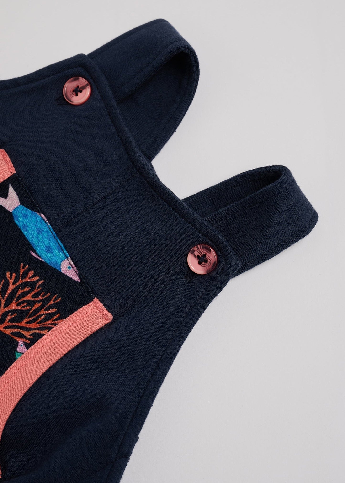 Close-up of adjustable straps set to short setting on navy short dungarees with vibrant fish print and coral buttons and coral trim.
