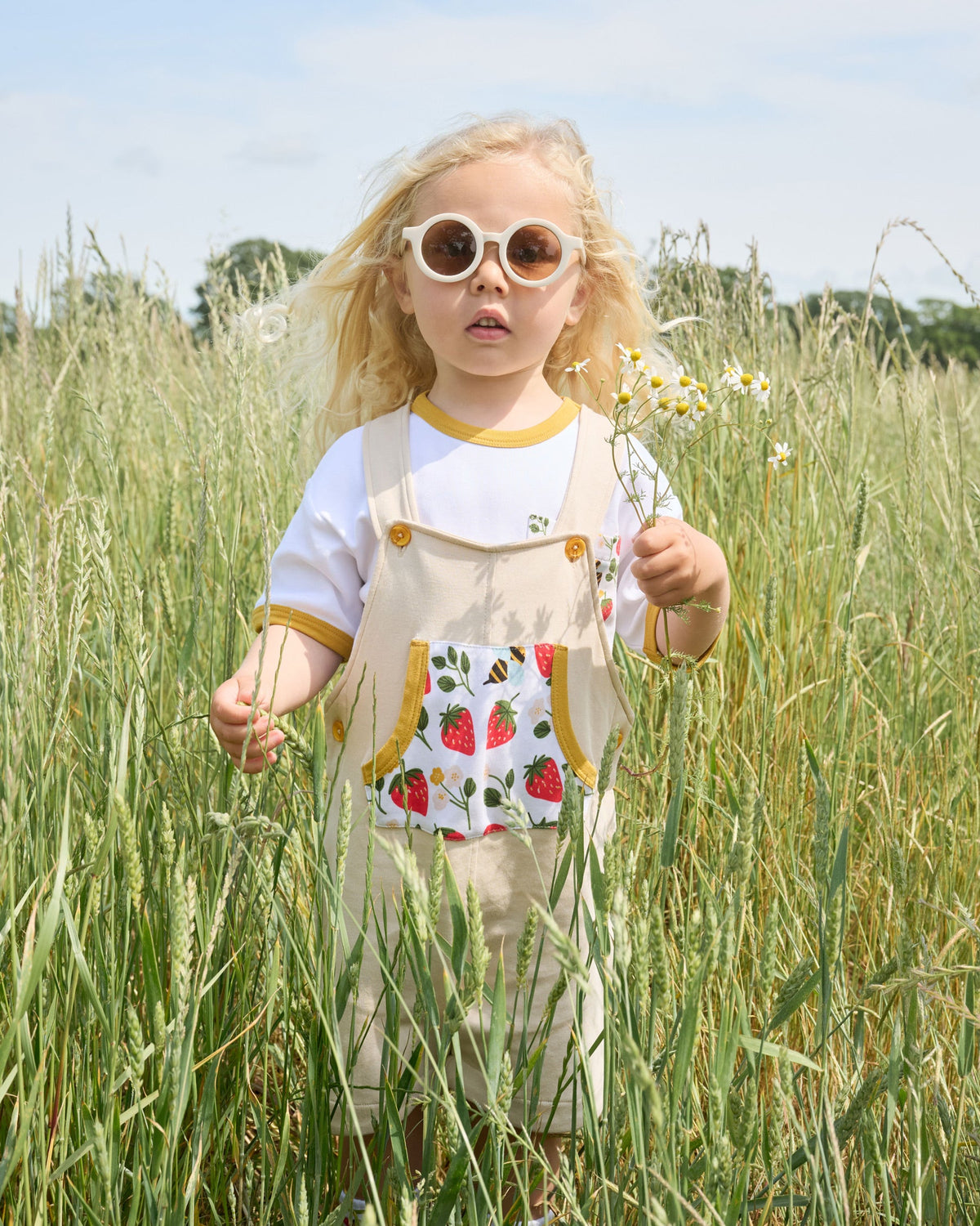 Young boy in a field wearing a white short sleeved t-shirt with fun strawberry and bees print square chest pocket and honey neck and sleeve trim under beige short dungarees.