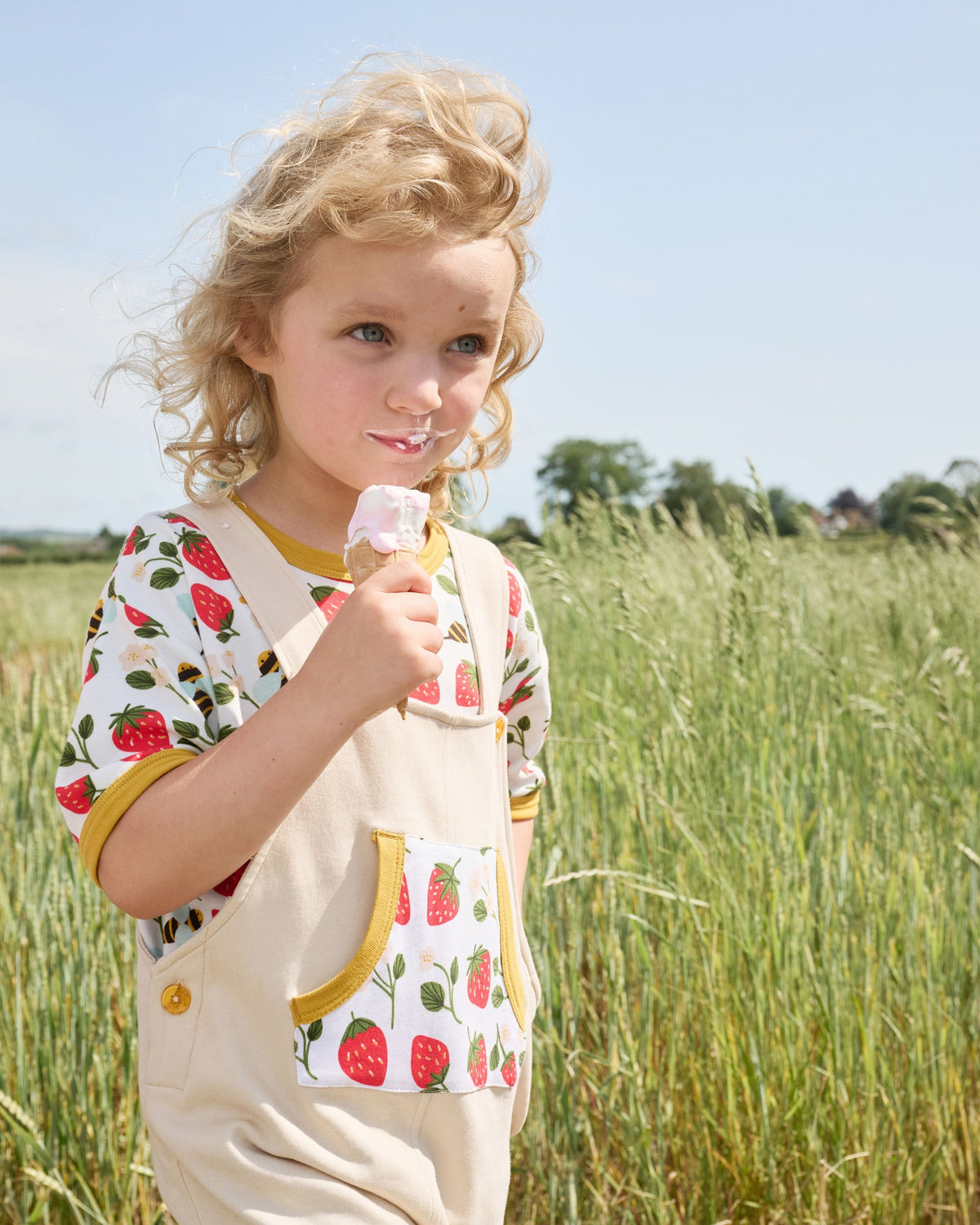 Little girl in a field eating ice cream wearing a short sleeved t-shirt featuring an all over strawberry and bees print with contrast honey neck and sleeve trim under a set of matching beige dungarees.