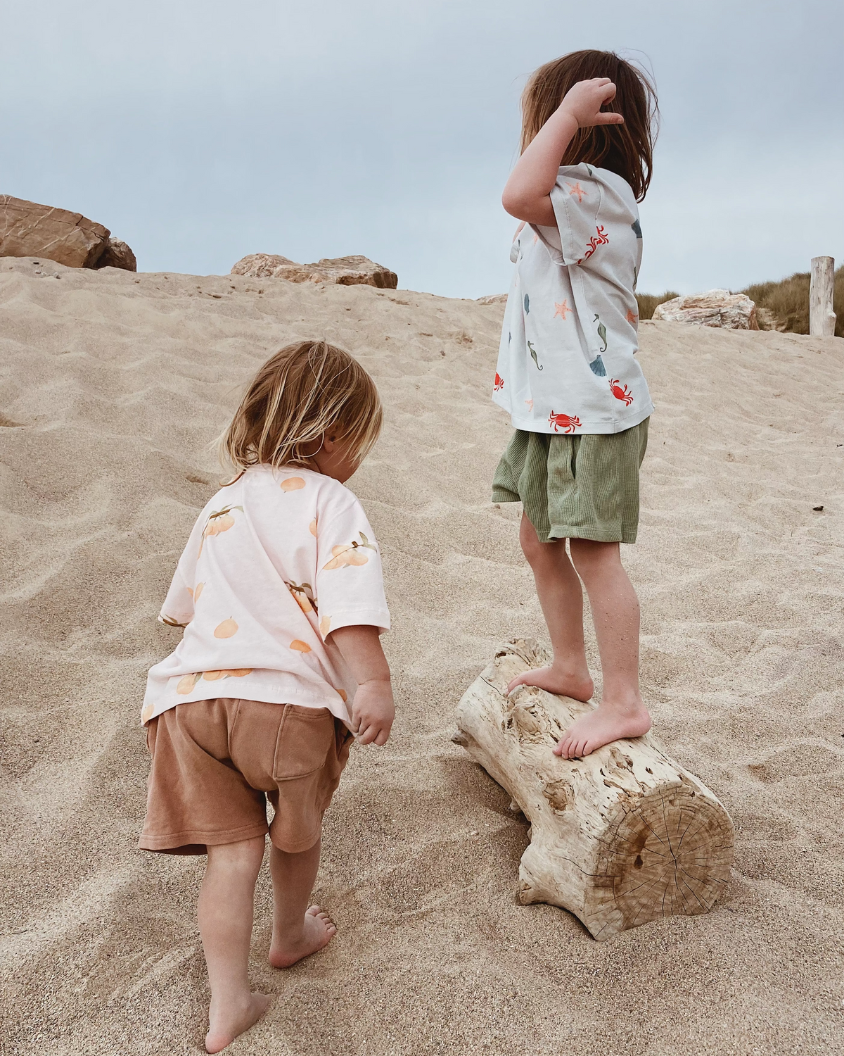 Children playing on a beach wearing Wildgreen organic cotton, gender-neutral t-shirts designed for sustainable, outdoor play. Ocean print and apricot peach pattern.