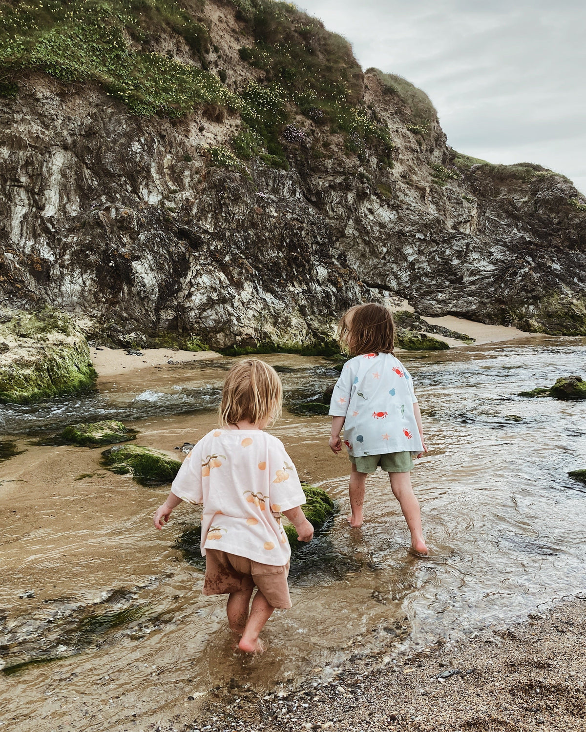 Two children playing in a beach river wearing Wildgreen organic cotton, gender-neutral t-shirts designed for sustainable, outdoor play.